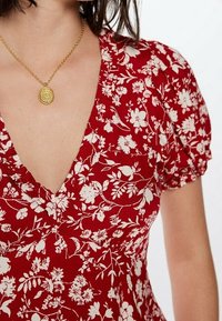 Rotes blumengemustertes Kleid mit tiefem V-Ausschnitt und Puffärmeln; strukturierter Stoff; kombiniert mit einer goldenen kreisförmigen Anhängerhalskette.