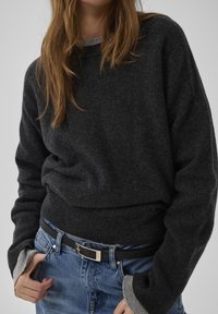 Dunkelgrauer Strickpullover mit langen Ärmeln, versehen mit einem gerippten Saum und Bündchen. Kombiniert mit hellblauem Denim und einem schwarzen Gürtel.