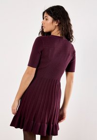 Giovane donna con lunghi capelli ricci, che indossa un vestito prugna a maniche corte con un orlo arricciato, in piedi davanti a uno sfondo semplice.