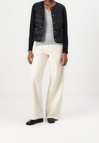 Zwarte puffervest met een gebreide mouw, grijze V-hals top en crème wijdlopende broek. Zwarte instappers vullen de outfit aan. Gladde texturen en subtiele details.