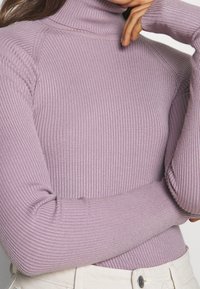 Kobieta w prążkowanym lawendowym swetrze z golfem i długimi rękawami, w połączeniu z jasnobeżowymi spodniami z widocznymi guzikami.