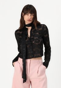 Black sheer blouse with floral embroidery, long sleeves, button closure, and a neck tie. Paired with high-waisted pink trousers.