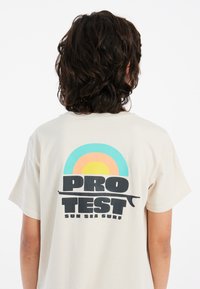 Kreminės spalvos trumparankovė marškinėliai, ant nugaros esantis grafinis piešinys: stilizuotas vaivorykštė turkio, oranžinės ir geltonos spalvų, su tekstu "PRO TEST".
