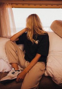 Czarna koszulka z krótkim rękawem z graficznym wzorem, beżowe dresy i białe sneakersy, siedzi na kanapie z białymi poduszkami i zasłonami.