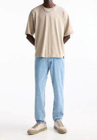 Camiseta de algodón beige con mangas cortas, combinada con jeans de mezclilla azul claro y zapatillas beige con detalles y cordones verdes.