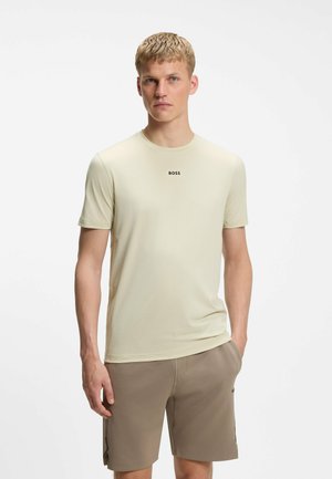 Ung mand med kort blondt hår iført en lys beige BOSS T-shirt og brune shorts, stående med den ene hånd i lommen.