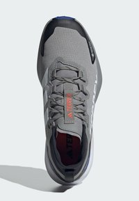 Scarpa sportiva grigia con tomaia in rete, dettagli neri, branding rosso e una suola bianca reattiva. Presenta punta rinforzata e chiusura con lacci.