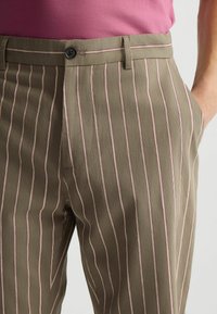 Olivgrüne gestreifte Hose mit schmalen weißen und rosafarbenen Streifen, ausgestattet mit einem Knopfverschluss und seitlichen Taschen. Texturiertes Material mit einer taillierten Passform.