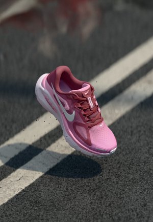 Ružová a bordová bežecká teniska Nike vo vzduchu nad asfaltom s bielymi pruhmi vozovky, vrhajúca tiene na zem.
