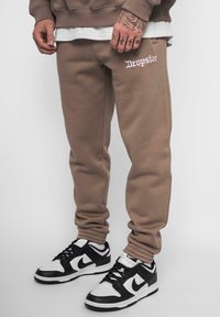 Braune Jogginghose mit elastischen Bündchen und weißem, gesticktem Text "Dropsize." Kombiniert mit schwarzen und weißen Nike-Sneakers. Weiches, strukturiertes Material.
