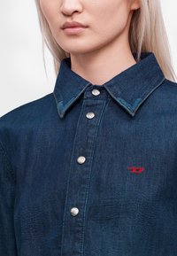 Gros plan d'une personne portant une chemise en denim bleu foncé avec des boutons en métal et un petit logo rouge brodé sur la poitrine.