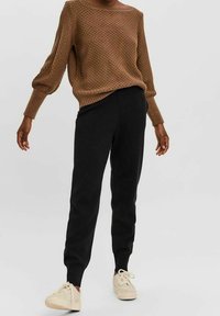 Personne portant un pull marron texturé, un pantalon de jogging noir slim et des baskets beige à lacets, debout devant un fond uni.