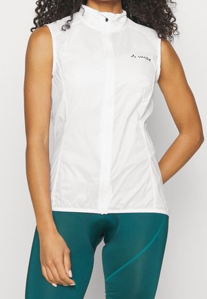 Gilet de cyclisme blanc sans manches avec fermeture éclair frontale porté sur un cuissard rembourré bleu sarcelle sur une personne debout.