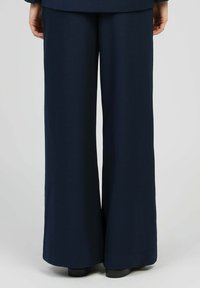 Personne portant un pantalon large bleu marine et des chaussures noires, debout avec les mains détendues le long du corps contre un fond clair.
