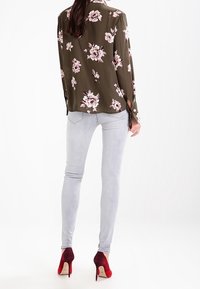 Blusa floral verde olivo de manga larga, combinada con jeans ajustados gris claro y tacones altos de gradiente rojo. Detalles de botones visibles en la blusa.
