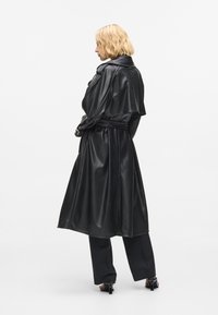 Μαύρο παλτό τύπου trench με ζώνη και υφή, με φαρδύ γιακά, άνετη εφαρμογή και μακριά μανίκια, συνδυασμένο με μαύρο παντελόνι.