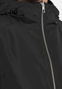 Schwarze leichte Jacke mit hohem Kragen, silbernem Reißverschluss und geraffter Kapuze. Glatte Textur mit minimalen Stichdetails.