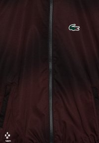 Veste légère bordeaux avec une texture en maille, dotée d'une fermeture éclair devant, de poches latérales et d'un logo crocodile vert sur la poitrine.