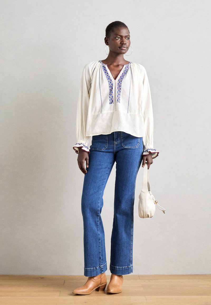 Blouse blanche à manches longues avec broderies bleues, associée à un jean décontracté bleu à pattes d'éléphant et des bottines beiges à la cheville. Le mannequin tient un petit sac à main blanc.