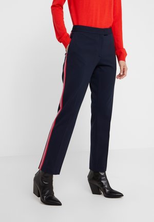 Personne portant un pantalon bleu marine avec des bandes rouges et blanches sur les côtés, un haut à manches longues rouge vif et des bottines noires à talons.