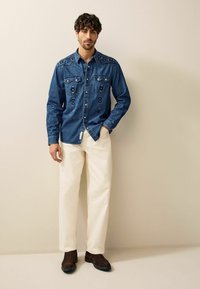 Camisa de denim con yugo en punta, costura decorativa y dos bolsillos en el pecho, combinada con pantalones color crema y botas de tobillo marrón oscuro.