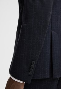 Donker marineblauwe blazer met een subtiel patroon, gestructureerde stof en vier zwarte knopen op de manchetten. Witte shirt zichtbaar eronder.