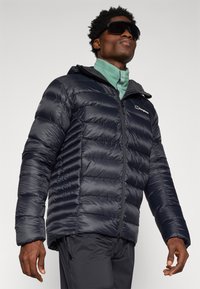 Jachetă puffer marină, cu design matlasat, închidere cu fermoar și glugă. Prezintă un finisaj ușor, strălucitor și un logo pe partea stângă a pieptului.