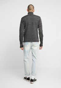 Tummanharmaa neulepusero, jossa on korkea kaula-aukko ja tekstuuriset kuviot, yhdistettynä vaaleansinihin käärittyihin farkkuihin ja mustiin tennareihin.