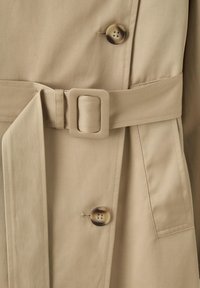 Trench-coat beige avec ceinture en tissu assortie et boucle carrée, avec deux boutons visibles brunâtres et une poche latérale.