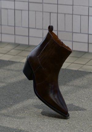 Sötétbarna bőr bokacsizma tömbsarkúval, hegyes orral és felhúzófüllel, strukturált járdán áll, csempézett fal mellett.