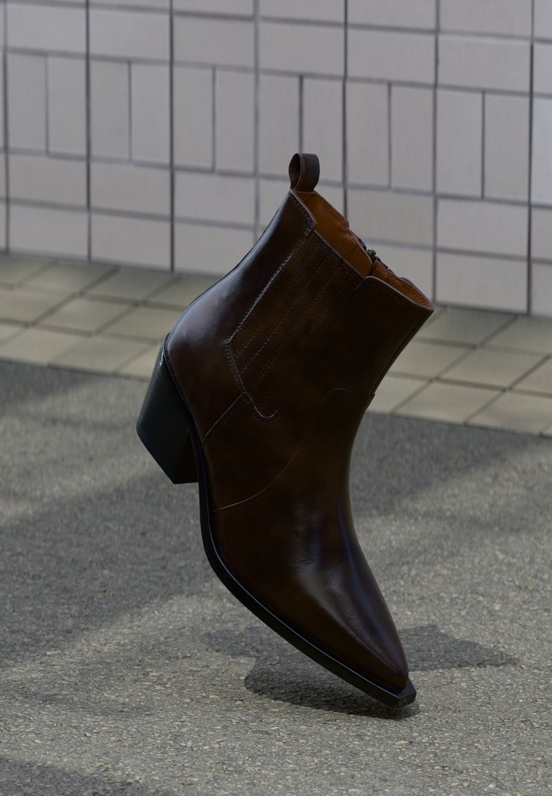 Σκούρο καφέ δερμάτινο μποτάκι αστραγάλου με χοντρό τακούνι, μυτερή μύτη και θηλιά, το οπ οίο στέκεται σε υφή πεζοδρομίου κοντά σε πλακωμένο τοίχο.