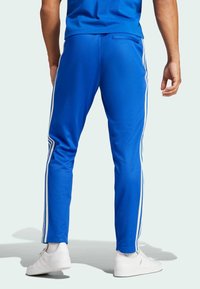 Man som bär blå träningsbyxor med vita ränder längs sidorna och vita sneakers, stående mot en ljus bakgrund.