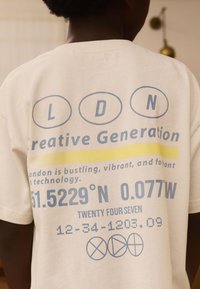 Flødefarvet bomulds-T-shirt med grafisk tekst i blå og gul. Indeholder koordinater og slogans i en moderne, digital skrifttype.
