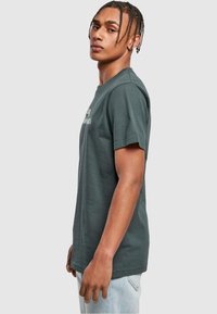 Mørkegrøn bomulds-t-shirt med korte ærmer, rund halsudskæring og tekstdesign foran. Modellen står sidelæns iført lyseblå jeans.