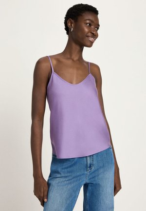 Woman smiling, wearing silver hoop earrings, a lavender sleeveless top with thin straps, and blue high-waisted jeans, standing against a plain background.