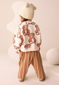 Vaalean beige huppari, jossa on karhupainatus, yhdistetty ruskeisiin raidallisiin housuihin. Asun viimeistelee kerma-valkoinen neulottu pipo, jossa on karhukorvat.