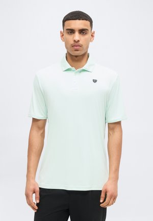 Νεαρός άνδρας που κοιτάει προς τα εμπρός φορώντας ανοιχτό πράσινο κοντομάνικο πόλο με μικρό λογότυπο ασπίδας στο στήθος και μαύρο παντελόνι.