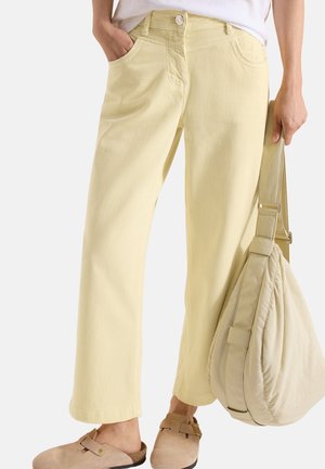 Kvinde iført lysegule bukser med lige ben og beige slip-on sko, der holder en matchende beige skuldertaske i den ene hånd.