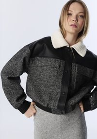 Jonge vrouw met handen op de heupen, gekleed in een grijze herringbone jas met een zwarte leren schouderstuk en een witte donzige kraag.