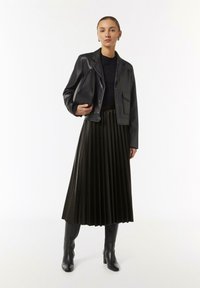Schwarze, cropped Lederjacke mit abgesetztem Kragen, kombiniert mit einem schwarzen, plissierten Midikleid und knielangen, schwarzen Stiefeln mit Absatz.