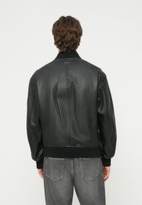 Vīrietis ar viļņainiem brūniem matiem, tērpies melnā ādas jakā un pelēkās džinsa biksēs, stāv ar muguru pret kameru uz baltas fona.