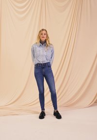 Camisa de mezclilla azul clara, suéter de cuello alto a rayas en blanco y negro, jeans oscuros ajustados y zapatos negros contra un suave fondo beige.