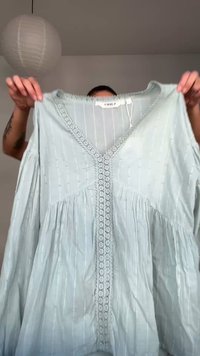 Persona che tiene in mano una blusa azzurro chiaro a maniche lunghe con righe verticali e rifiniture in pizzo lungo lo scollo a V e la parte anteriore.