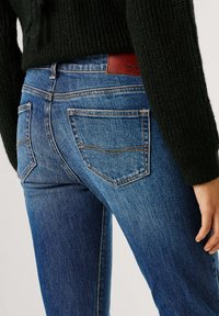 Sinised teksapüksid, millel on liibuv lõige, tagataskud ja kontrastne õmblusliin. Vöökohal on pruun nahast silt.