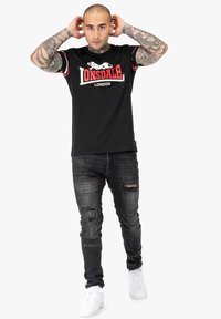 T-shirt nera a girocollo con dettagli rossi e bianchi, con logo "LONSDALE LONDON". Abbinata a jeans grigi strappati e sneakers bianche.