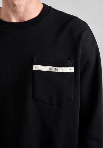 Mustmustelane särk ümmarguse kaelusega ja ristkülikukujulise rinnakotiga. Kotil on valge horisontaalne riba, millel on tekst "MOSCHINO." Pehme kangast tekstuur.