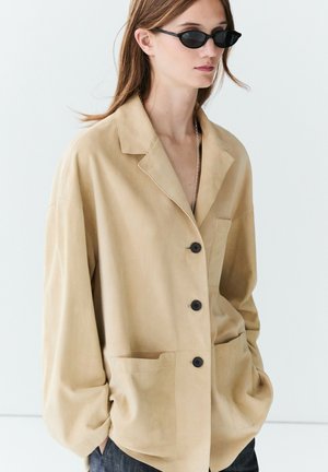 Frau trägt beige übergroße, geknöpfte Jacke mit vorderen Taschen und schwarze schmale ovale Sonnenbrille, Hände in den Taschen, vor einem einfarbigen hellen Hintergrund.