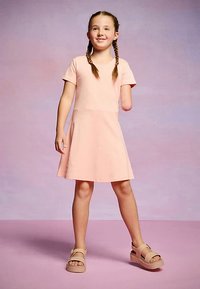 Šviesiai rožinė trumpomis rankovėmis suklostyta suknelė su platėjančia sijono dalimi, lygiu audiniu ir V formos iškirpte, derinama su rudu sandalais, stovinti prie pastelinio fono.