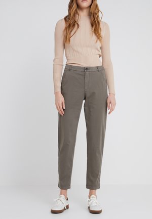 Vrouw staand, draagt een beige geribde lange mouw top, olijfgroene hoge taille broek en witte sneakers met bruine zolen.