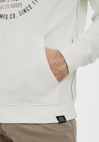 Hvid sweatshirt med frontlomme, sort syning og tekstlogo. Har ribbede manchetter og kant, samt et lille mærke nederst.